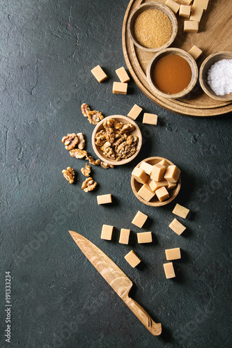 Salted caramel fudge candy served on wooden board with fleur de sel, caramel sauce, brown cane sugar and caramelized walnuts in wood bowls over black texture background. Top view, space. Dessert set
