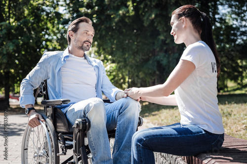 Sympathy and support. Positive nice young woman looking at the disabled man and holding his hand while showing her support