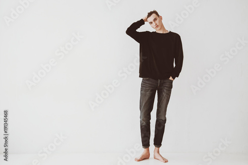 A young man, a student stands against a white wall in the room, dressed in short jeans and a black sweater, barefoot. The guy is a model.