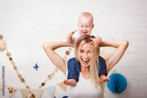 A cute handsome one-year-old baby sitting by her mother's neck grabs her by the hair and laughs. A beautiful young mother with her son play laughing and look at the camera. Mothers Day. Close-up