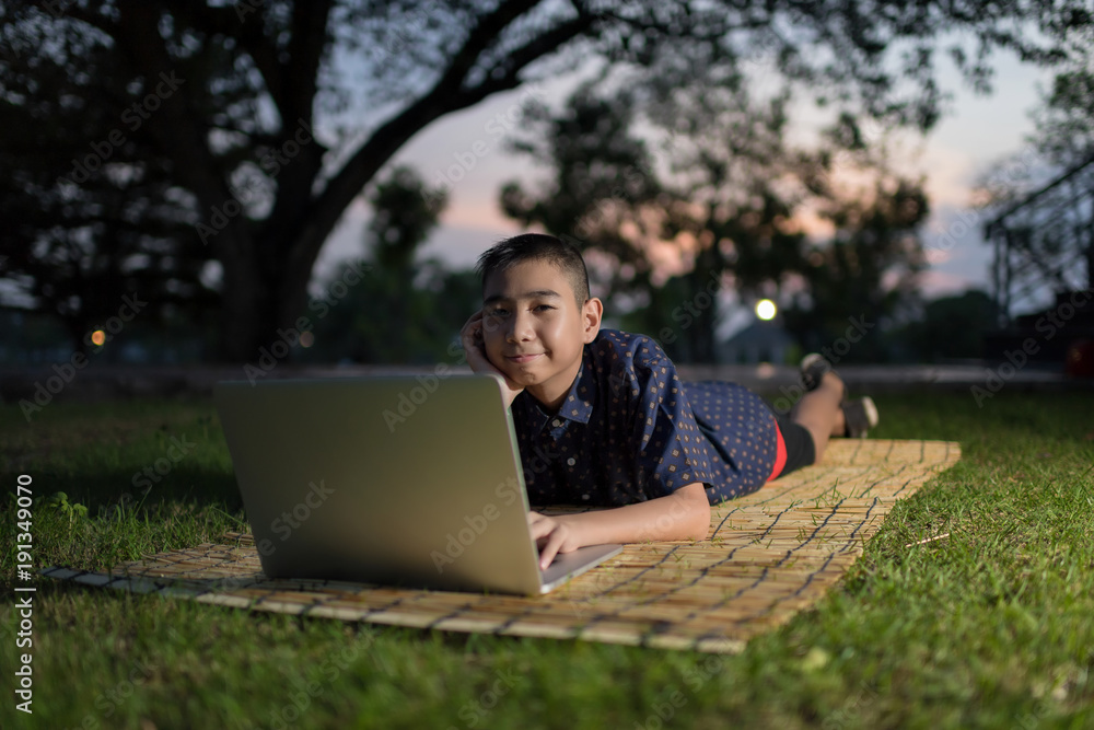 Beautiful boy sitting down on green grass in a park and using a laptop while studying and doing his homework in evening lighting Beautiful boy sitting down on green grass in a park and using a laptop while studying and doing his homework in evening lighting