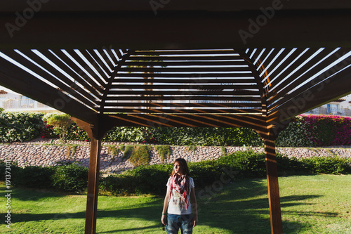 Portrait of young attractive beautiful cutie brunette woman standing in summer warm sunny day near green lawn under wooden arch and roof. Sensuality, shadows. Concept of tourism, vacation, recreation.