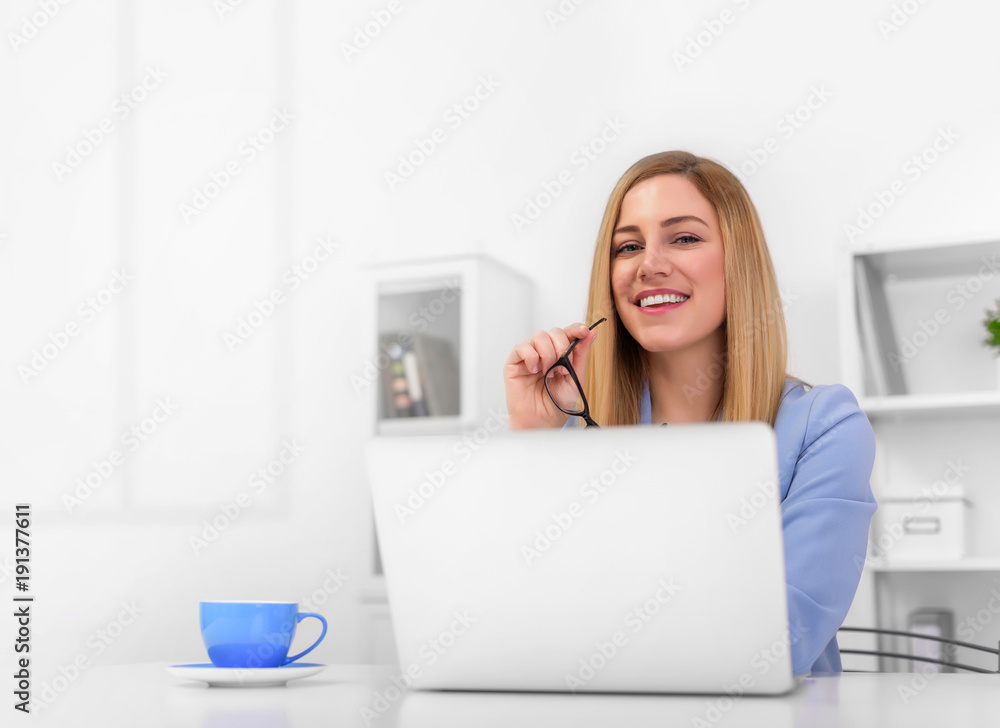 Attractive blonde in a blue suit is sitting at the office desk with a laptop and glasses in her hands. Attractive blonde in a blue suit is sitting at the office desk with a laptop and glasses in her hands.