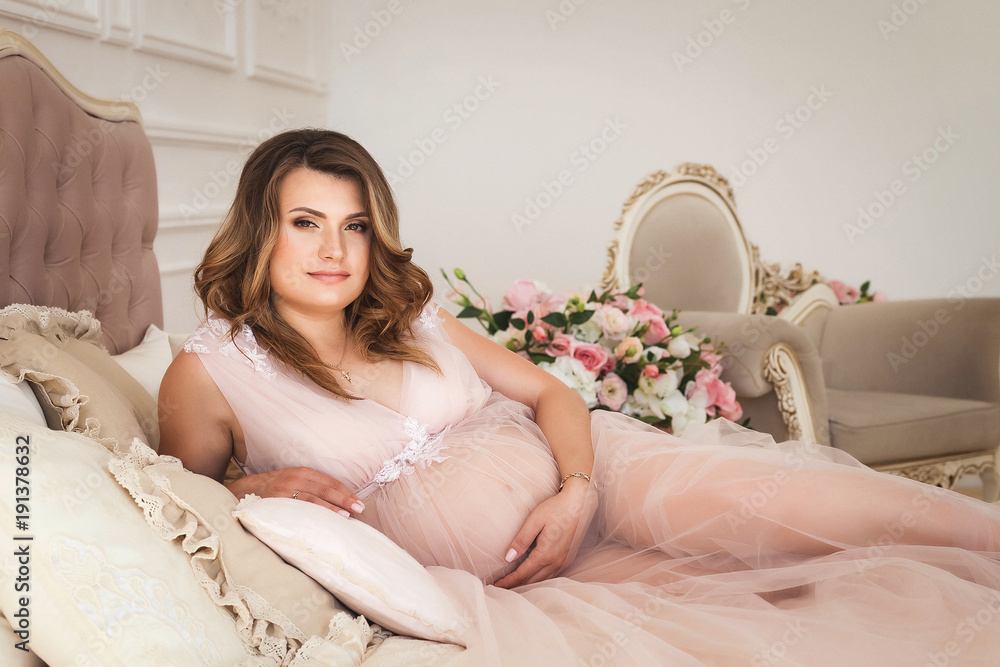 Young pregnant woman with curly hair in long elegant dress lying in cozy bed in luxury room the morning and hugging her belly Young pregnant woman with curly hair in long elegant dress lying in cozy bed in luxury room the morning and hugging her belly