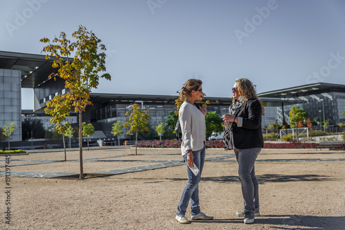 Two mature women wearing casual clothes, talking in the middle of a large park, with an office building in the background