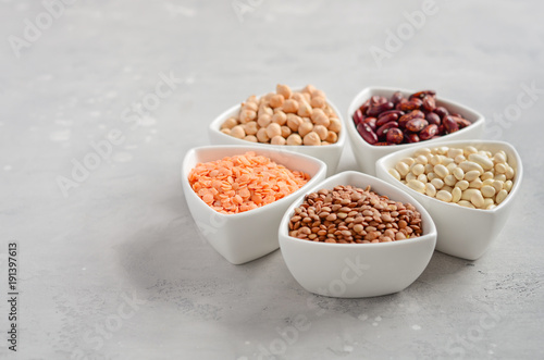 Selection of dry legumes, lentils and peas in white bowls on gray concrete background. Selective focus, copy space.