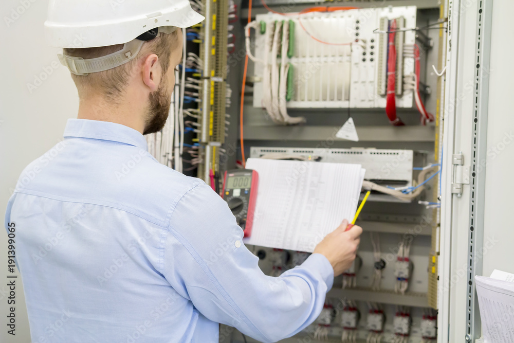 Engineer in white helmet reads design drawing against electric industrial panel. Service worker analyzes the electrical circuit in electrical automation control box with controllers. Engineer in white helmet reads design drawing against electric industrial panel. Service worker analyzes the electrical circuit in electrical automation control box with controllers.