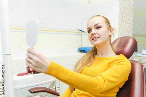 Young woman checking her teeth at mirror after dental treatment in stomatology clinic