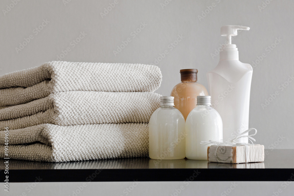 Pile of towels, bottles with shampoo, body lotion, shower milk and handmade soap on neutral background. - obrazy, fototapety, plakaty 