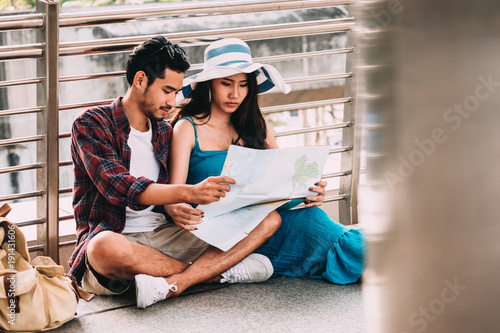 Couple Asian travelers are sitting on city street and looking at interesting places to travel from paper map. Vacation Concept