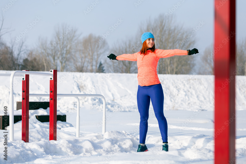 A young woman in a bright blue hat, orange sweater and elk smiles and does a warm-up before training on the run on a sports field on a bright winter day