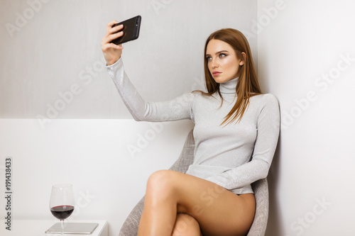 Portrait of beautiful lady in sweater sitting in chair and taking photo on frontal cellphone camera with glass of red wine near isolated
