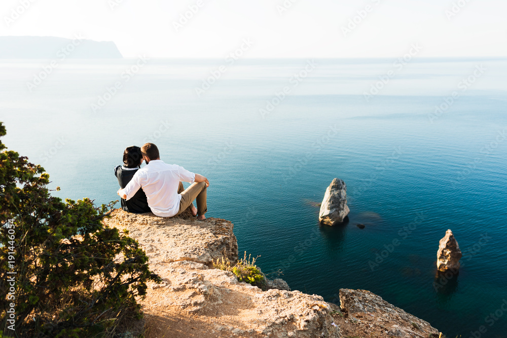 Man and woman sitting on the edge of a cliff by the sea. Honeymoon ...