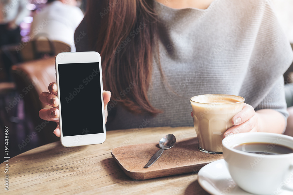 Mockup image of a woman holding and showing white mobile phone with blank black desktop screen with coffee cups on vintage wooden table Mockup image of a woman holding and showing white mobile phone with blank black desktop screen with coffee cups on vintage wooden table