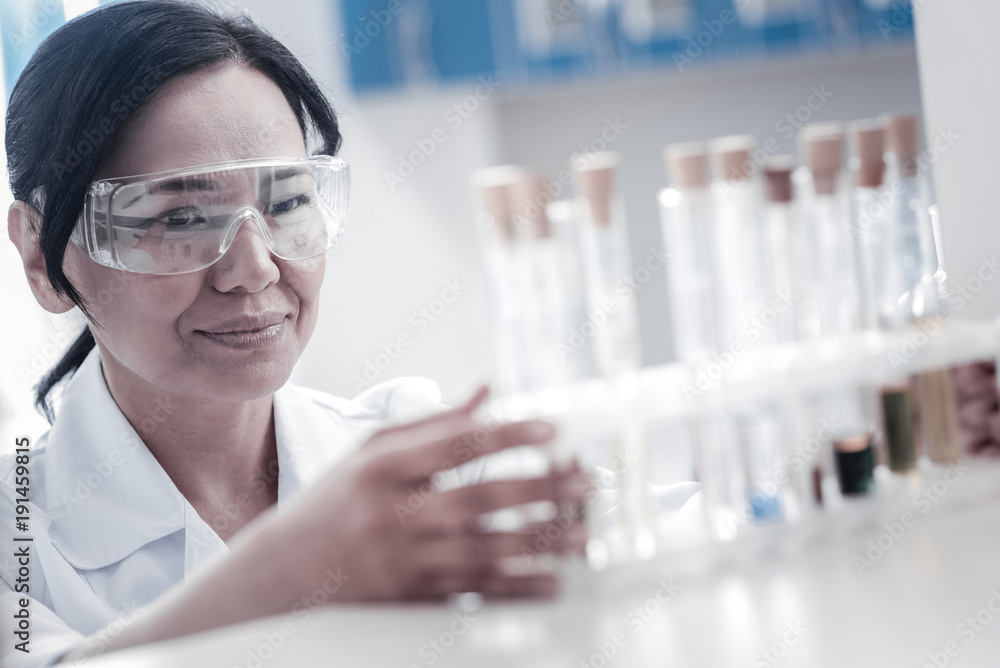 Innovative studies. Selective focus on a positive minded mature lady in safety glasses smiling while looking at a set of test tubes containing different substances. Innovative studies. Selective focus on a positive minded mature lady in safety glasses smiling while looking at a set of test tubes containing different substances.