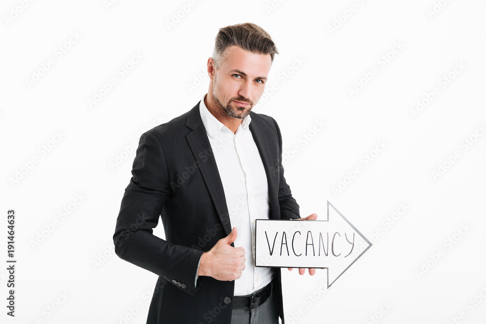 Image of confident office worker in black jacket holding speech arrow pointer with word vacancy in hand and showing thumb up, isolated over white wall Image of confident office worker in black jacket holding speech arrow pointer with word vacancy in hand and showing thumb up, isolated over white wall