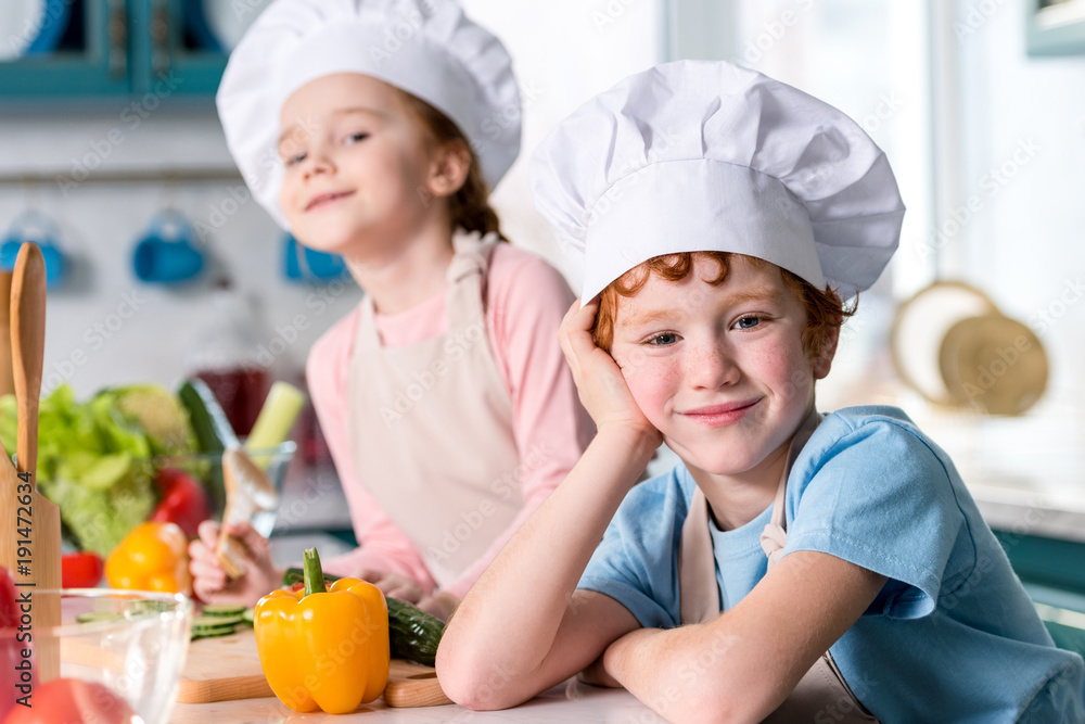 adorable siblings in chef hats and aprons smiling at camera while cooking in kitchen adorable siblings in chef hats and aprons smiling at camera while cooking in kitchen
