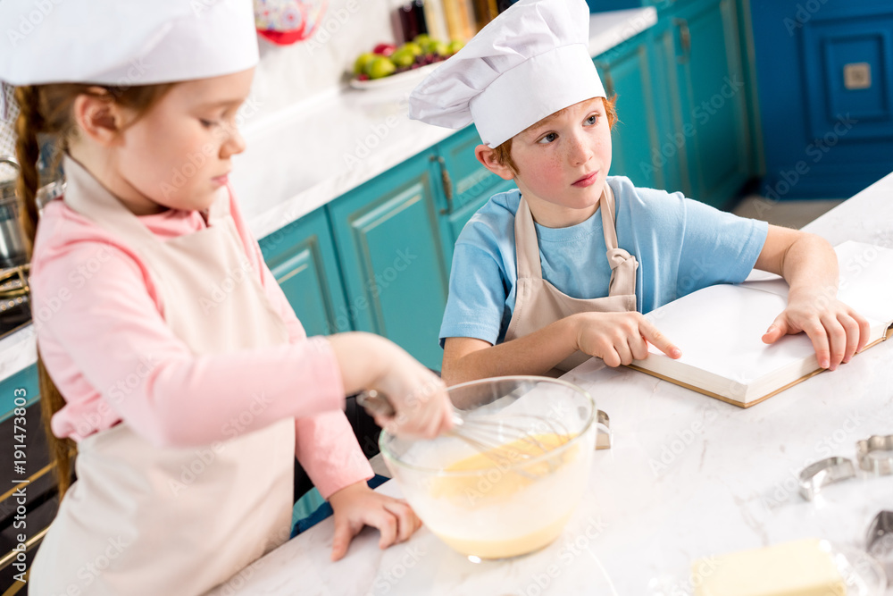 cute little kids in chef hats making dough and reading cookbook in kitchen cute little kids in chef hats making dough and reading cookbook in kitchen