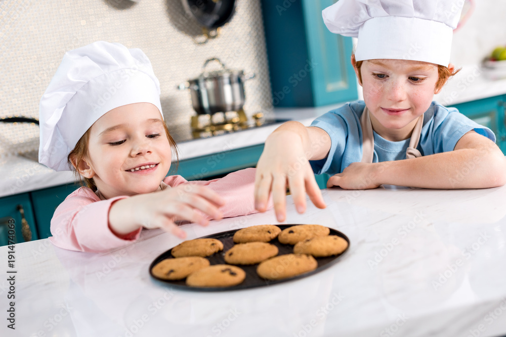 happy little kids in chef hats eating tasty cookies in kitchen happy little kids in chef hats eating tasty cookies in kitchen