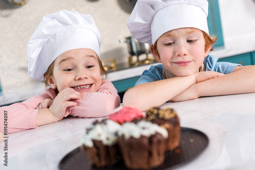happy kids in chef hats looking at delicious cupcakes on foreground happy kids in chef hats looking at delicious cupcakes on foreground