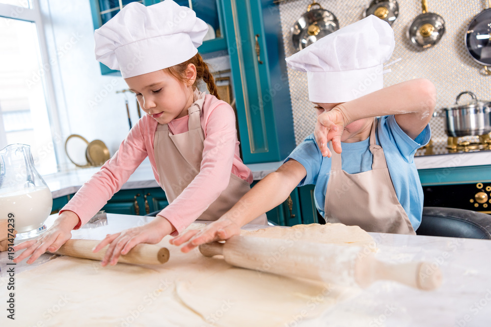 focused little children in chef hats and aprons rolling dough in kitchen focused little children in chef hats and aprons rolling dough in kitchen