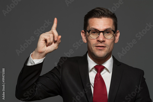 Portrait of young positive businessman wearing glasses and formal suit pointing his finger up against gray background