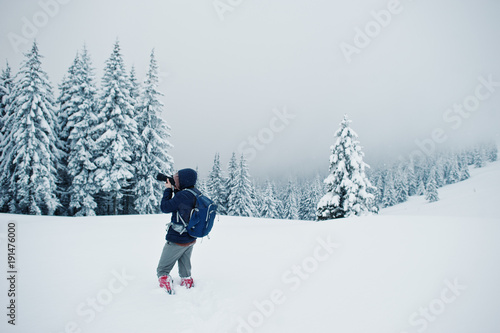 Man tourist photographer with backpack, at mountain with pine trees covered by snow. Beautiful winter landscapes. Frost nature.