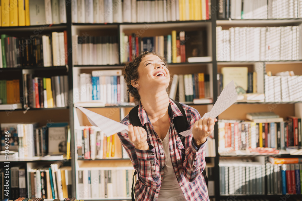 Joyful smiling student girl holding papers and celebrating while standing in the library. Joyful smiling student girl holding papers and celebrating while standing in the library.