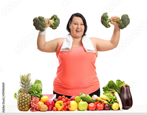 Overweight woman exercising with broccoli dumbbells behind a table with fruit and vegetables
