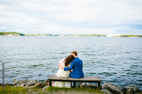 Newlyweds are relaxing on a bench and sitting before a river. Bride and groom is resting on nature