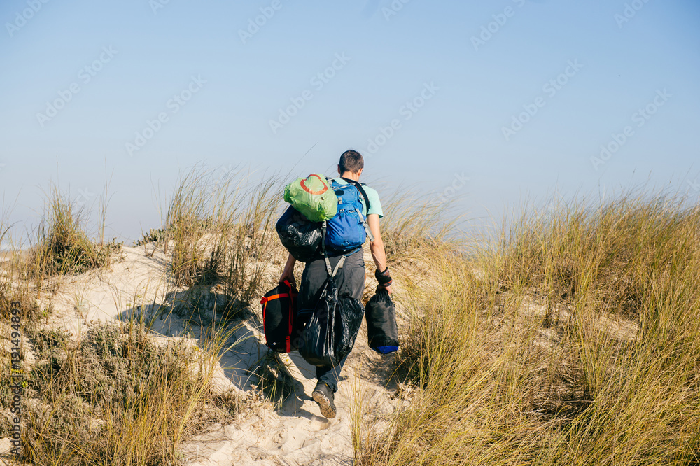 Homeless hermit carrying many bags with belongings in sandy dunes beyond sea in sunny day. Man from behind travelling alone. Unknown male with backpacks and heavy buggage in hands hiking outdoor.