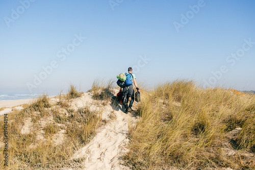 Homeless hermit carrying many bags with belongings in sandy dunes beyond sea in sunny day. Man from behind travelling alone. Unknown male with backpacks and heavy buggage in hands hiking outdoor.