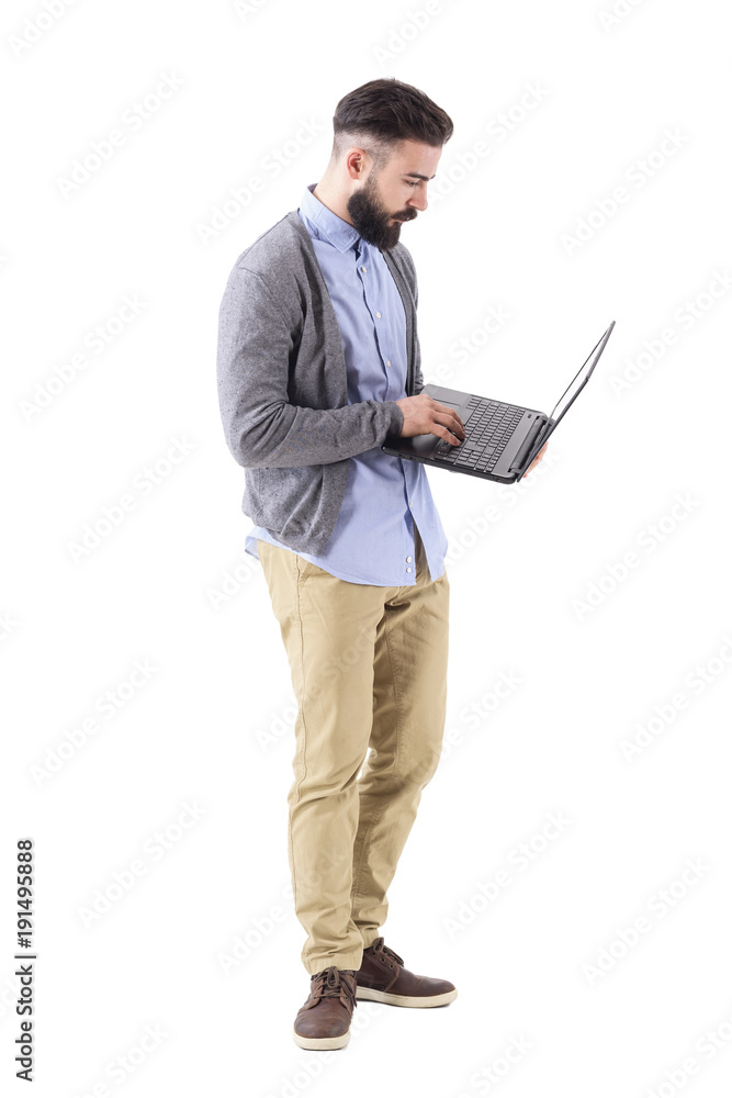 Side view of stylish bearded man typing on laptop keyboard looking at computer. Full body length portrait isolated on white studio background. Side view of stylish bearded man typing on laptop keyboard looking at computer. Full body length portrait isolated on white studio background.