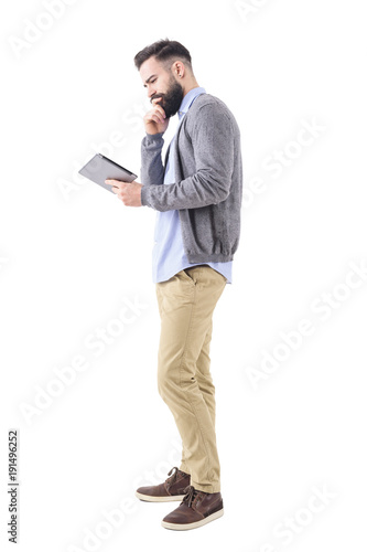 Thinking young business man looking at tablet with hand on chin. Full body length portrait isolated on white studio background. 