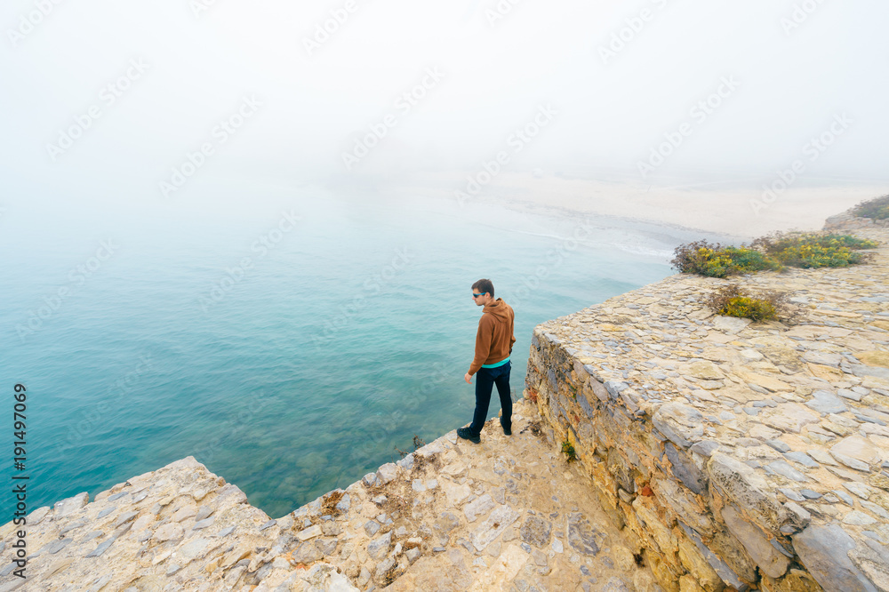 Man at edge of the world. Traveler male tourist standing beyond african fortress tower with fog and mist on ocean background. Young boy with camera at big height in Peniche in Portugal. Tranquillity.