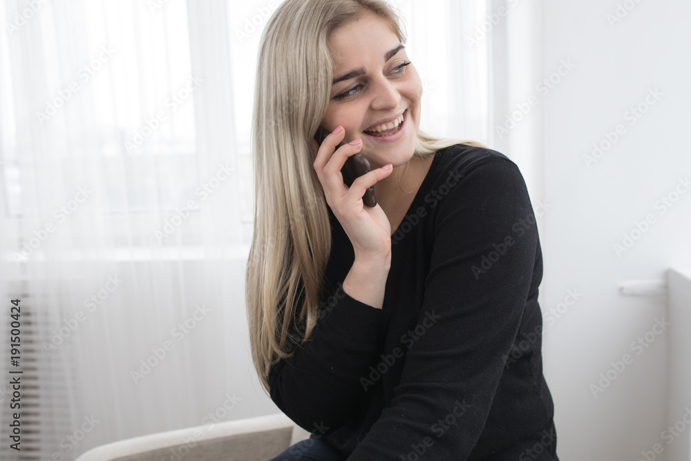 white blonde girl sitting on a white armchairon a white background in black pullover and posing and smiling with her smartphone talking with friends white blonde girl sitting on a white armchairon a white background in black pullover and posing and smiling with her smartphone talking with friends