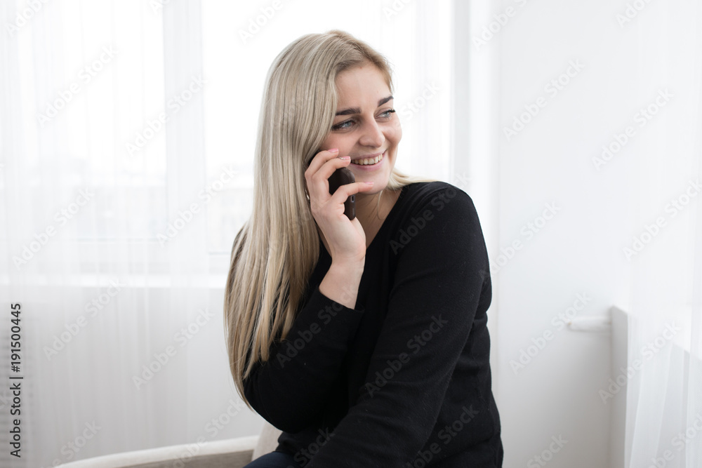 white blonde girl sitting on a white armchairon a white background in black pullover and posing and smiling with her smartphone talking with friends white blonde girl sitting on a white armchairon a white background in black pullover and posing and smiling with her smartphone talking with friends