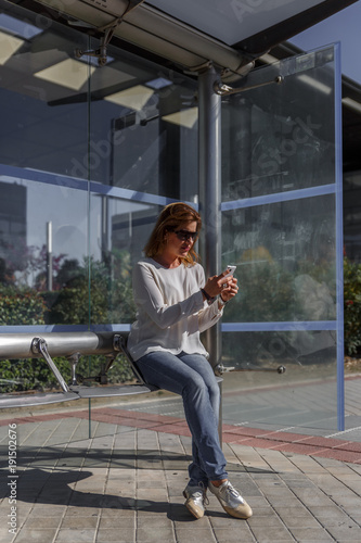 Business woman in casual clothes carefully reviews something important on her mobile phone, while waiting sitting at the bus stop, on a sunny day
