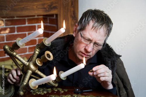 A rich greedy elderly man looks at coins under the light of a burning candle. The collector looks at his wealth with lit candles.