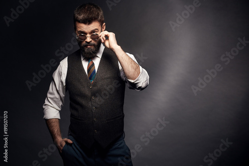 Studio portrait of attractive handsome man with dark hair, mustache and beard in white shirt, brown vest and colorful tie takes off round sunglasses