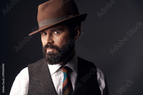 Studio portrait of attractive handsome man with dark eyes, dark hair, mustache and beard in white shirt, brown vest, colorful tie and brown classic hat.