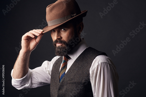 Studio portrait of handsome man with dark hair, mustache and beard in white shirt, brown vest, colorful tie, holding brown hat on head. Old fashioned style.