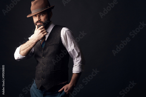 Studio portrait of handsome man with dark hair, mustache and beard with hand on it in white shirt, brown vest, colorful tie, brown hat on head. Old fashioned style.