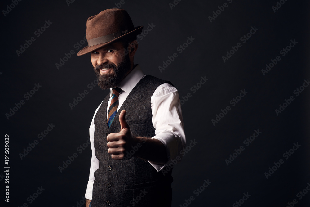 Studio portrait of smiling handsome man with dark hair and mustache in white shirt, brown vest, colorful tie, brown hat with thumb up. Old fashioned style, gentleman. Studio portrait of smiling handsome man with dark hair and mustache in white shirt, brown vest, colorful tie, brown hat with thumb up. Old fashioned style, gentleman.
