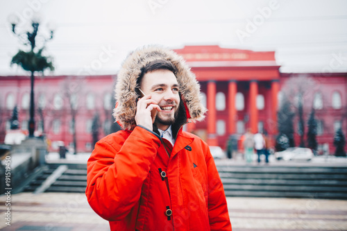 young guy with beard and red jacket in hood a student uses mobile phone, holds in his hand near the head, talking on the phone with smile on the background of red building the university or college