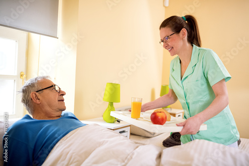 Bilde på lerret Young smiling nurse brings breakfast to senior patient