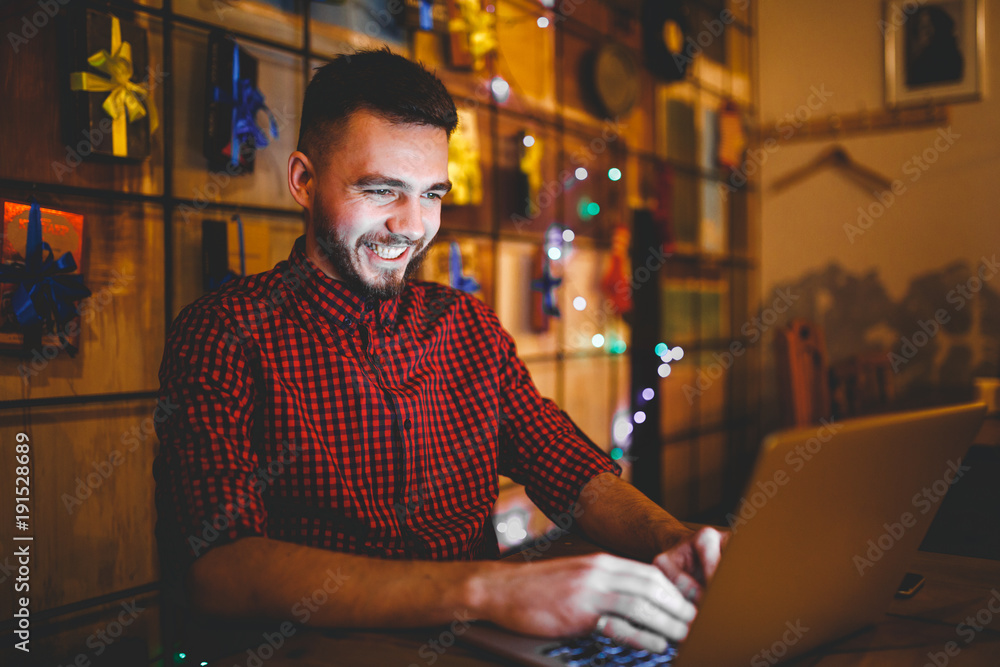Theme training and computers. A young man with a beard and in a shirt uses a laptop, prints on the keyboard in a coffee shop at a wooden table in the evening. Christmas decor and hang a garland Theme training and computers. A young man with a beard and in a shirt uses a laptop, prints on the keyboard in a coffee shop at a wooden table in the evening. Christmas decor and hang a garland