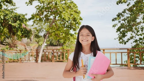 A charming philippine schoolgirl with a backpack and books in a park off the coast. A girl joyfully poses, raising her hands up with textbooks in her hands. Warm sunny day. photo