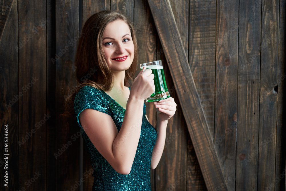 Obraz premium Sexy smiling woman holding mug of green beer for St. Patricks Day celebrations on wooden background, copy space. Girl in emerald sequins dress holding glass of beer in pub. Greeting card, March 17