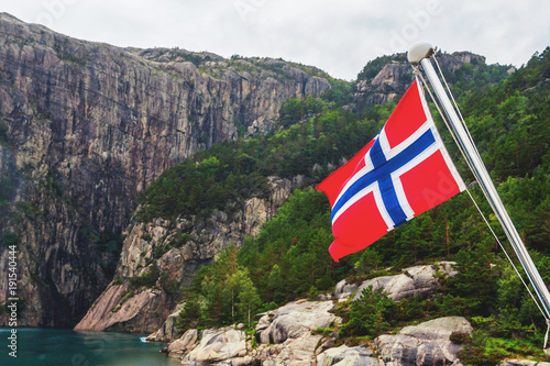 Norwegian flag on the fjord surrounded by mountains. Cloudy and rainy weather, dramatic sky.